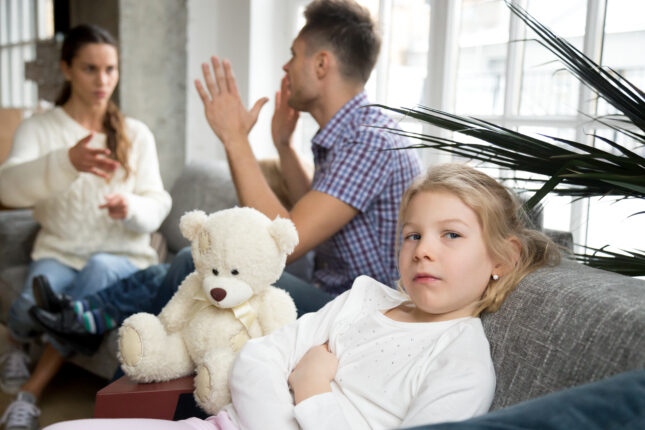 Trauriges kleines Mädchen mit Teddybär, während Eltern im Hintergrund streiten