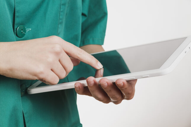 Eine Pflegekraft in grüner Arbeitskleidung bedient ein Tablet. Der Fokus liegt auf den Händen, während die Person mit einem Finger den Bildschirm berührt. Das Tablet wird horizontal gehalten und symbolisiert den Einsatz digitaler Technologien im Gesundheitswesen