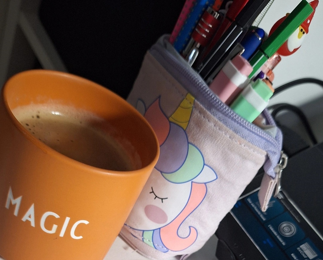 Eine orange Tasse mit der Aufschrift 'MAGIC', ein Einhorn-Federmäppchen und ein Drucker auf einem Schreibtisch.