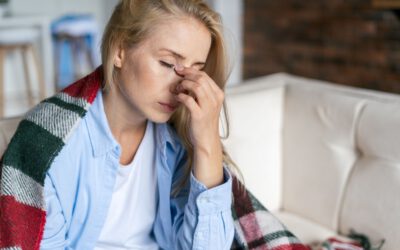 Eine erschöpfte Frau mit blonden Haaren sitzt auf einem Sofa, hält sich die Nasenwurzel und sieht müde oder gestresst aus. Sie trägt ein blaues Hemd und hat eine warme karierte Decke um die Schultern gelegt. Der Hintergrund zeigt eine gemütliche Wohnzimmersituation mit unscharfen Details.