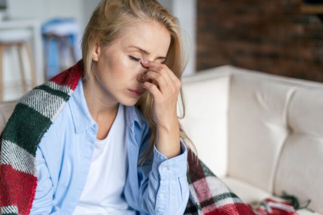 Eine erschöpfte Frau mit blonden Haaren sitzt auf einem Sofa, hält sich die Nasenwurzel und sieht müde oder gestresst aus. Sie trägt ein blaues Hemd und hat eine warme karierte Decke um die Schultern gelegt. Der Hintergrund zeigt eine gemütliche Wohnzimmersituation mit unscharfen Details.