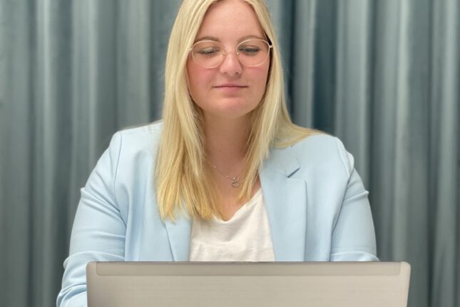 Frau arbeitet konzentriert am Laptop im Büro, mit Tasse und Smartphone auf dem Tisch.