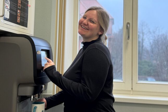Lächelnde Asklepios-Mitarbeitende steht in einem hellen Pausenraum am Kaffeevollautomaten und hält eine Tasse.